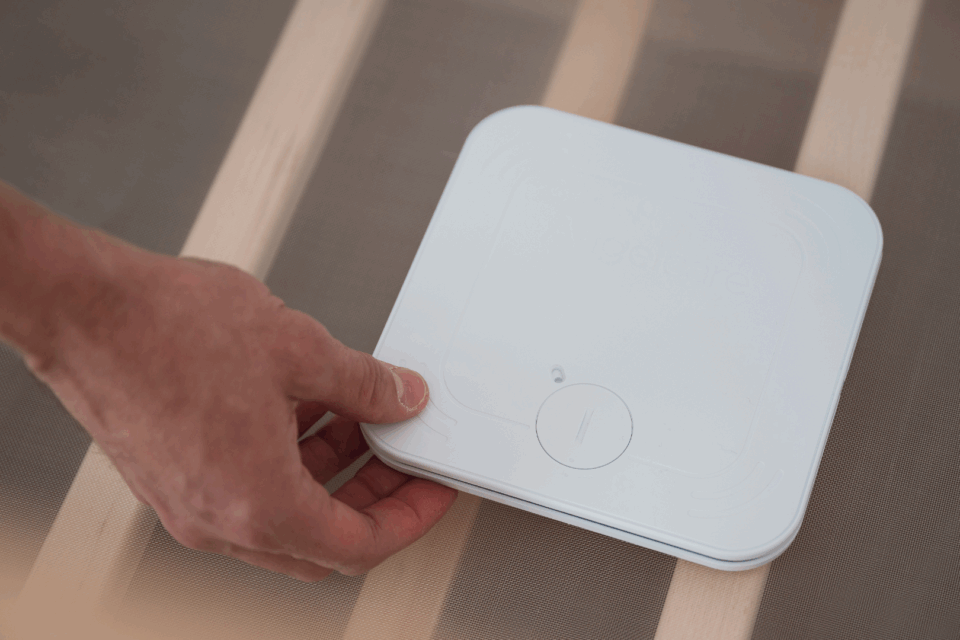 A hand is placing a monitor beneath a baby mattress in a cot