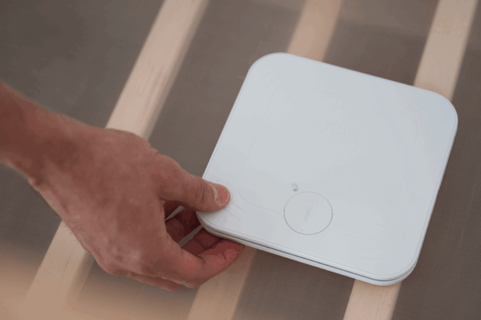 A hand is placing a monitor beneath a baby mattress in a cot