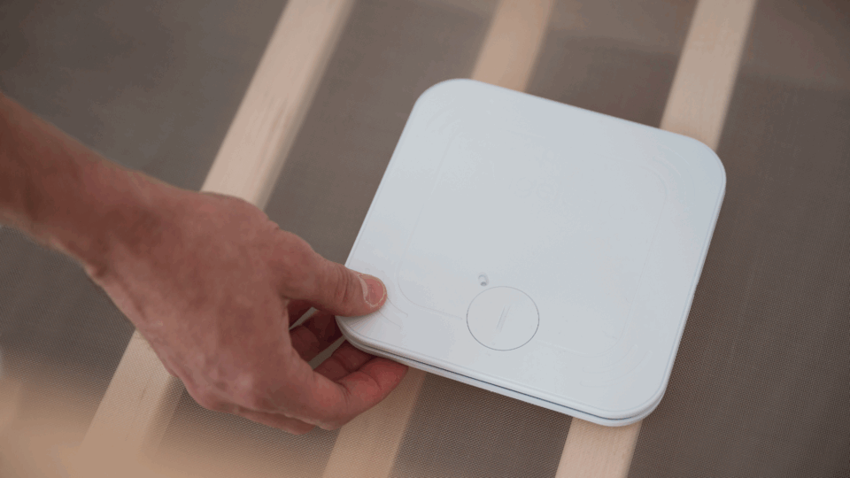 A hand is placing a monitor beneath a baby mattress in a cot