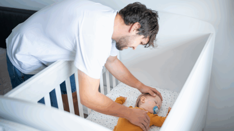 Dad checking on baby sleeping in their cot with a dummy in.
