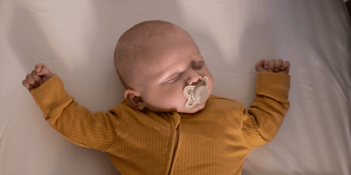 A baby is sleeping with their arms either side of their head. They're wearing yellow, have light skin and a dummy in their mouth.