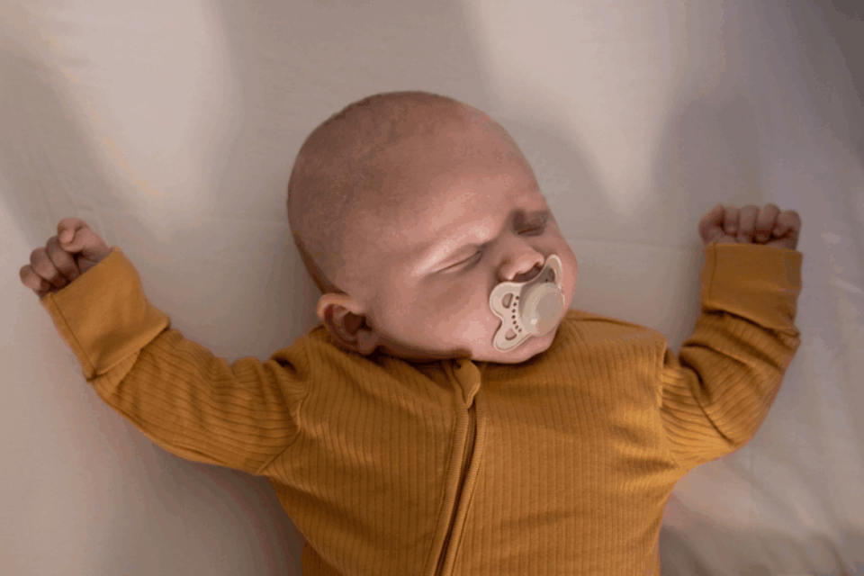 A baby is sleeping with their arms either side of their head. They're wearing yellow, have light skin and a dummy in their mouth.