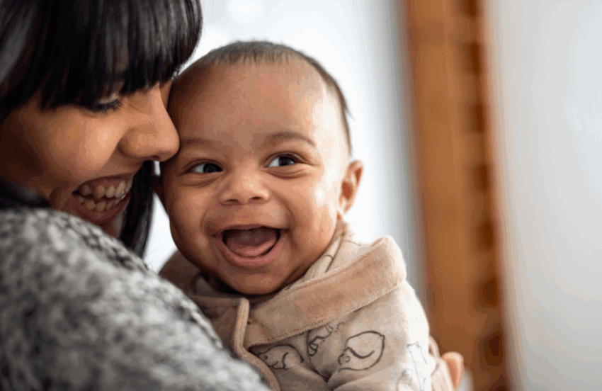 A baby is looking to their side with a huge, wide open grin. They have light to medium skin and the mum is looking down at them and smiling, with her face closely pressed to the baby.