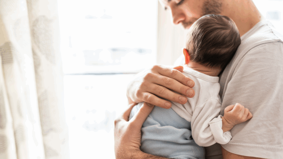 A dad is holding their baby on their chest, with his hand on the baby's back.