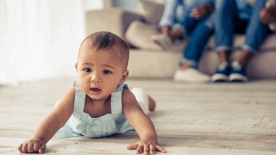 Baby is doing tummy time on the floor of the living room. They have medium skin and wear a light blue vest. They are looking intriguingly at something.