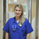 Emily, wearing scrubs and a stethoscope, standing in a hospital corridor smiling at the camera.