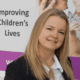 Georgina wearing a shirt and jacket, smiling at the camera. She had blond hair, and is standing in front of a backdrop that says 'Improving children's lives'.