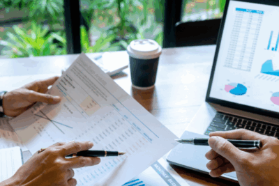 A close up of a laptop with graphs, and paper with data. Two pairs of hands are inspecting the data with a pen.