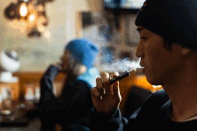 A man is facing side on, vaping in what looks to be a cafe. He is wearing a beanie hat.