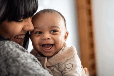 A baby is looking to their side with a huge, wide open grin. They have light to medium skin and the mum is looking down at them and smiling, with her face closely pressed to the baby.