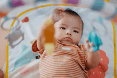 A baby is laying on their back playing with toys dangling above. They are wearing an orange and white stripey baby grow.