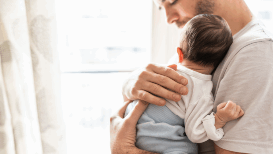 A dad is holding their baby on their chest, with his hand on the baby's back.