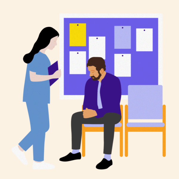 Man sitting on a chair in a hospital corridor in front of a notice board. A health professional is standing beside him talking to him.