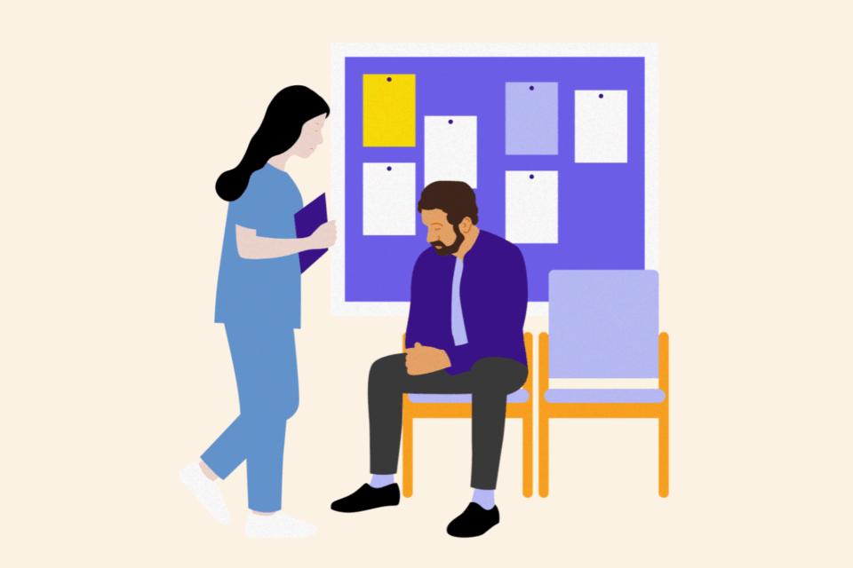 Man sitting on a chair in a hospital corridor in front of a notice board. A health professional is standing beside him talking to him.