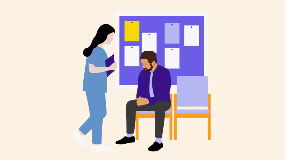 Man sitting on a chair in a hospital corridor in front of a notice board. A health professional is standing beside him talking to him.