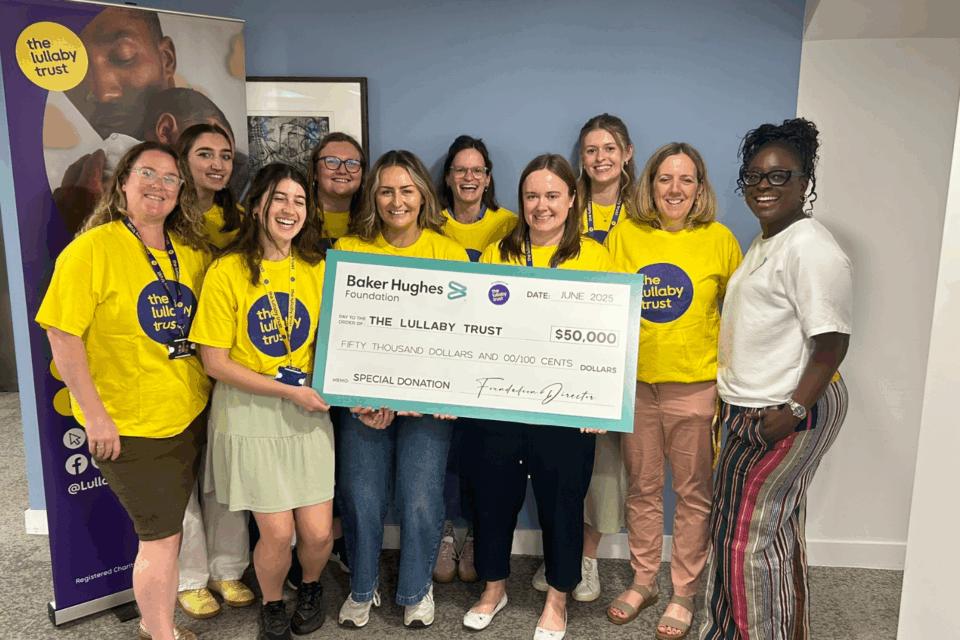 Seyi and lots of Lullaby Trust staff all in yellow t-shirts, smiling at the camera holding a big cheque for $50,000.