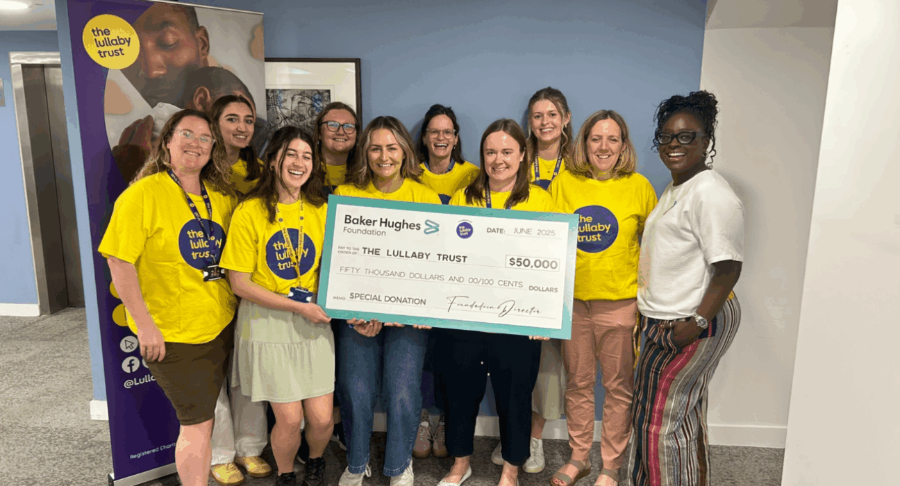 Seyi and lots of Lullaby Trust staff all in yellow t-shirts, smiling at the camera holding a big cheque for $50,000.