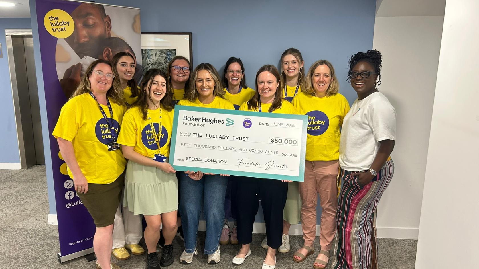 Seyi and lots of Lullaby Trust staff all in yellow t-shirts, smiling at the camera holding a big cheque for $50,000.