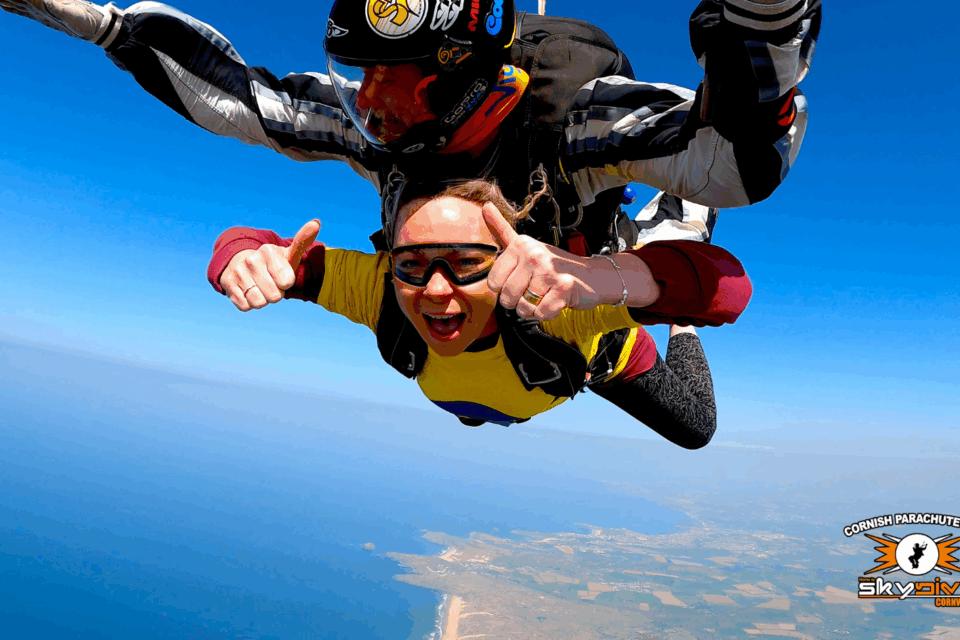 Katie strapped to an instructor, in the middle of her skydive giving thumbs up to the camera.