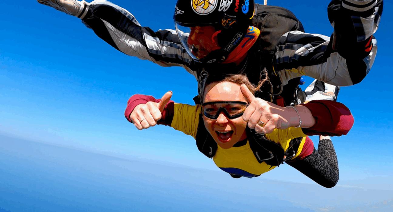 Katie strapped to an instructor, in the middle of her skydive giving thumbs up to the camera.