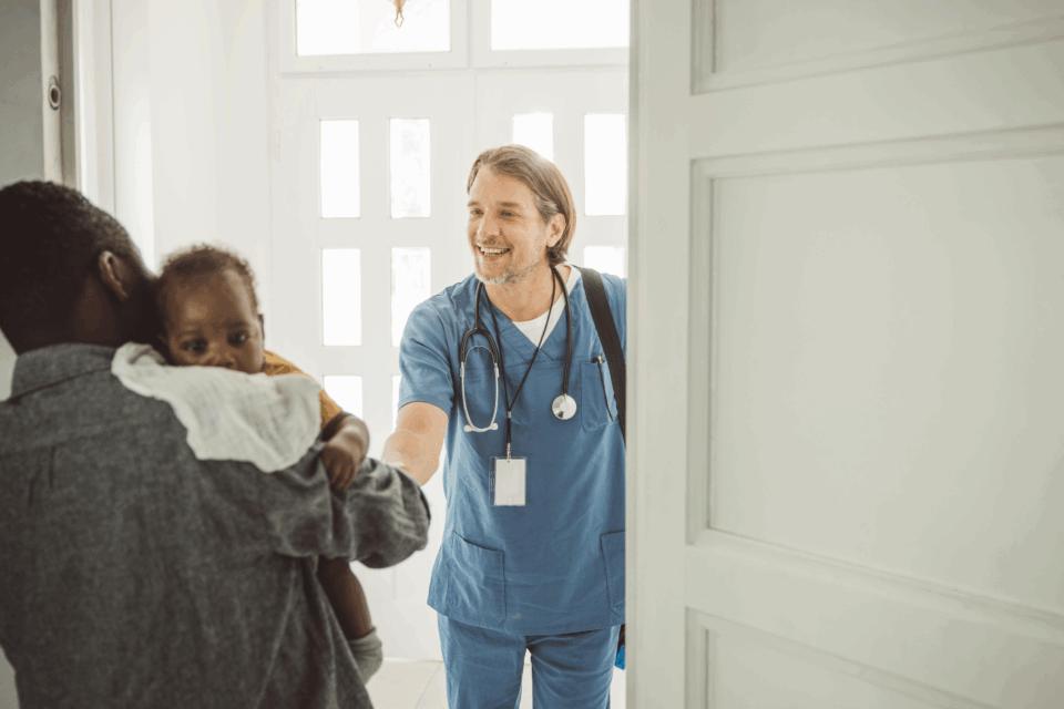 Health professional beside the front door of a home, shaking hands with a father who is holding a baby.