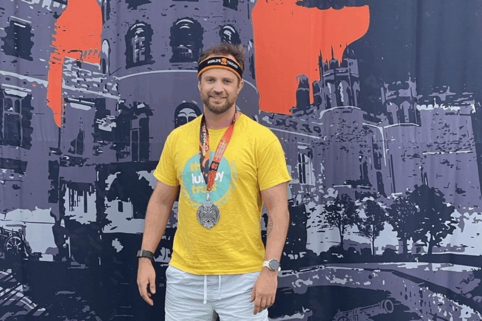 Stefan wearing a Lullaby Trust t-shirt and a medal, standing in front of a big tough mudder backdrop.