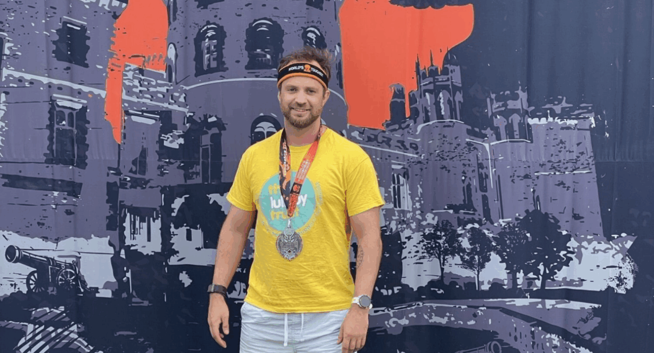 Stefan wearing a Lullaby Trust t-shirt and a medal, standing in front of a big tough mudder backdrop.