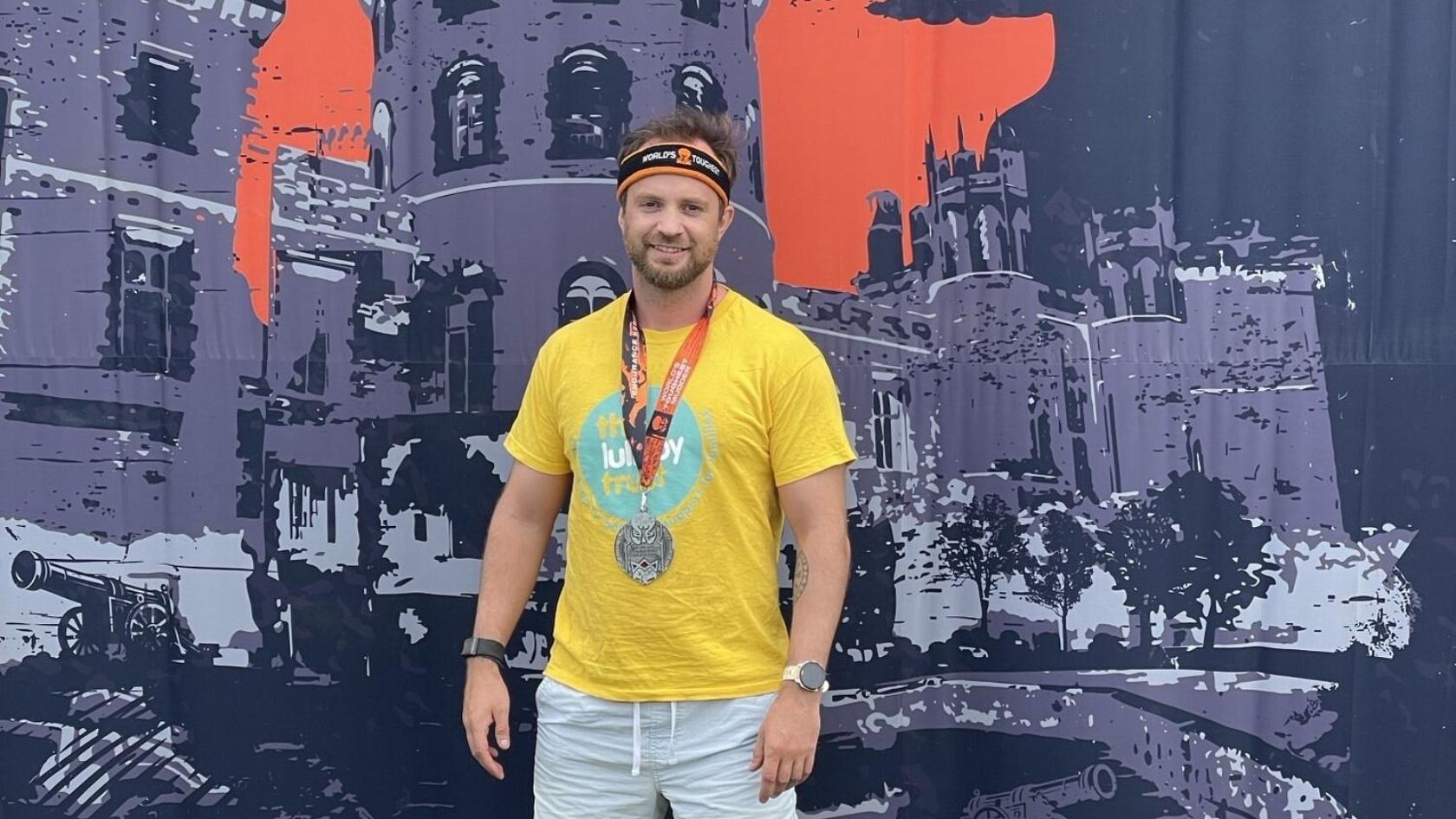 Stefan wearing a Lullaby Trust t-shirt and a medal, standing in front of a big tough mudder backdrop.