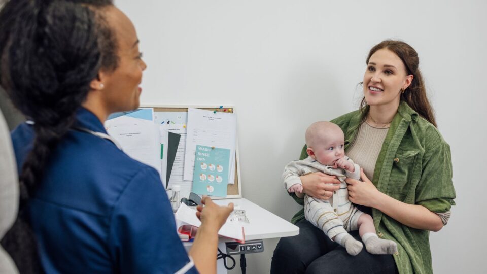 A mother is talking to a nurse at an appointment. They are smiling and the mother holds a young baby in her arms.