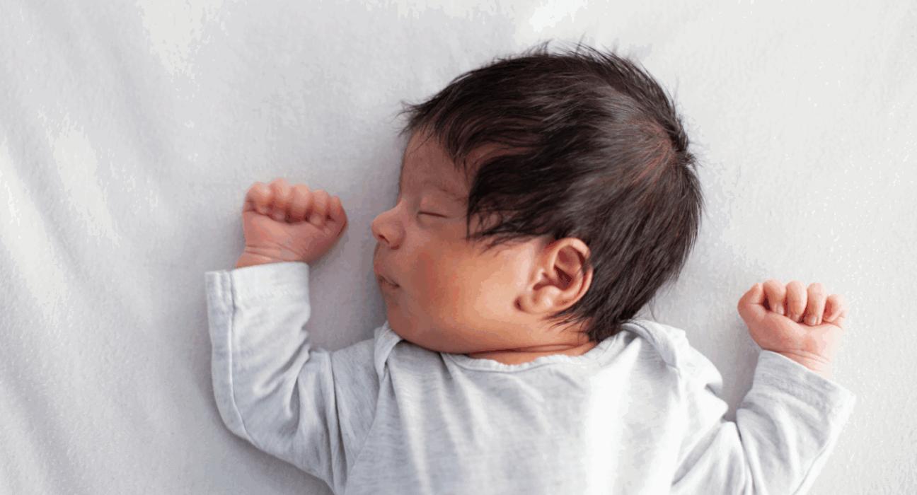 A baby is sleeping in a safer sleep position. They are on their back with a clear cot around them. They have dark hair and medium skin, and their arms are held up at either side of their head. They are sleeping.