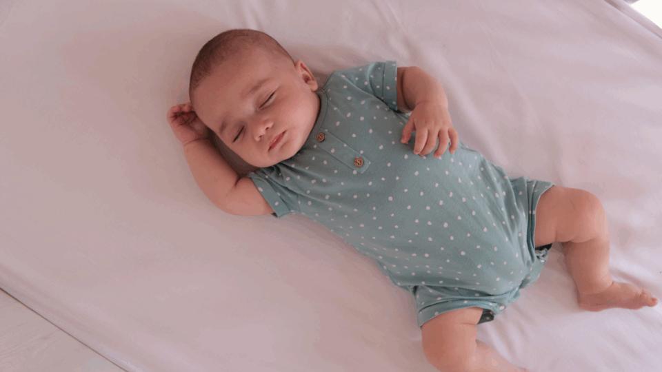 A baby sleeping safely on their back in an empty cot. They have one arm by their head, and another on their stomach. They are wearing a blue, polka-dot baby-grow.