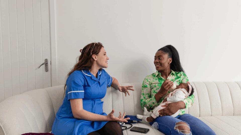 Mother bottle-feeding a baby, sitting on a sofa beside a health professional who is talking to her.