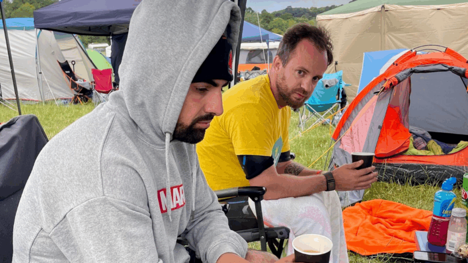 Stefan wearing a Lullaby T-shirt, sat on a chair next to a friend, with a tent next to them.