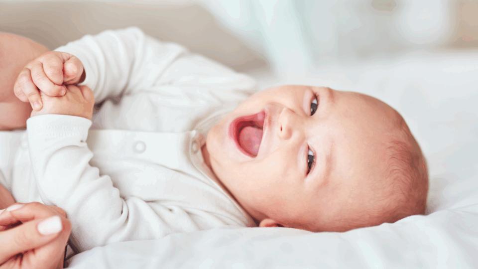 Baby is laying down smiling and giggling, grasping their hands together. They have light skin and wear a white babygrow.