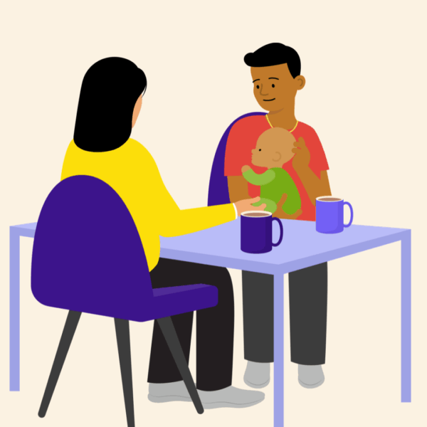 A man holding a baby, sitting at a table opposite another person. There are mugs on the table.
