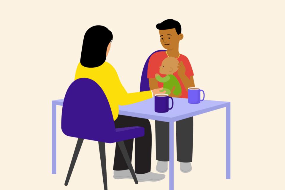 A man holding a baby, sitting at a table opposite another person. There are mugs on the table.
