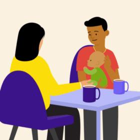 A man holding a baby, sitting at a table opposite another person. There are mugs on the table.