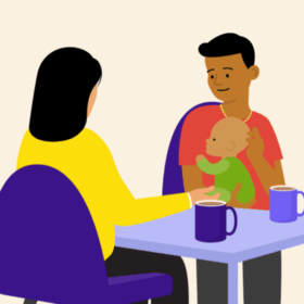 A man holding a baby, sitting at a table opposite another person. There are mugs on the table.