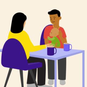 A man holding a baby, sitting at a table opposite another person. There are mugs on the table.