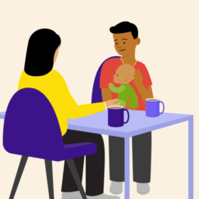 A man holding a baby, sitting at a table opposite another person. There are mugs on the table.