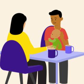 A man holding a baby, sitting at a table opposite another person. There are mugs on the table.