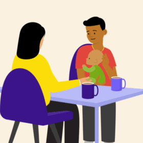 A man holding a baby, sitting at a table opposite another person. There are mugs on the table.