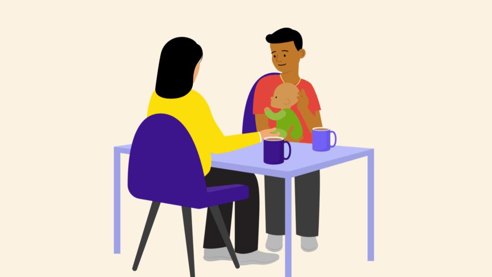 A man holding a baby, sitting at a table opposite another person. There are mugs on the table.