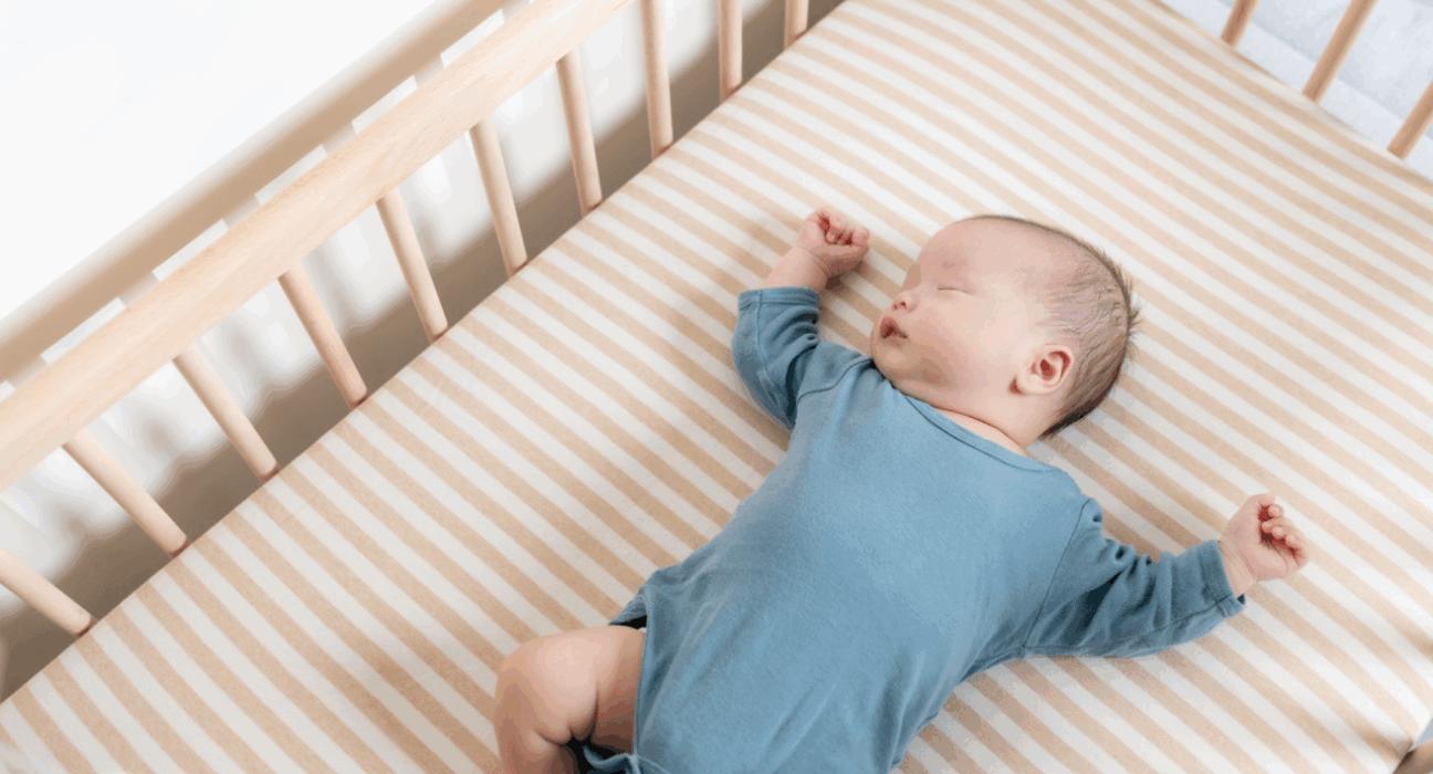 A baby in a blue baby grow is sleeping on their back in a wooden cot with a firm mattress.