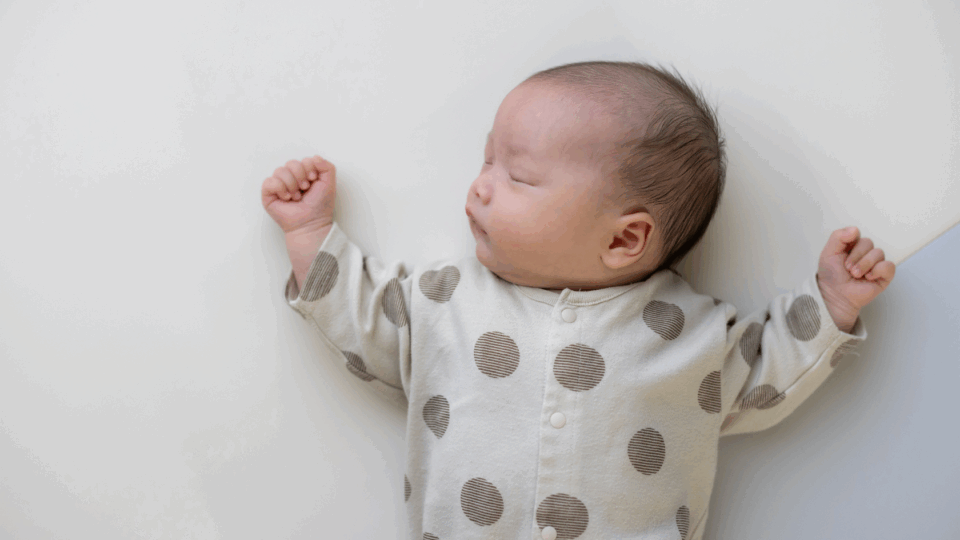 A baby is sleeping in on their back, with their head tilted to the side. They have dark hair, light skin and wear a polkadot baby grow. Their arms are rested either side of their head.