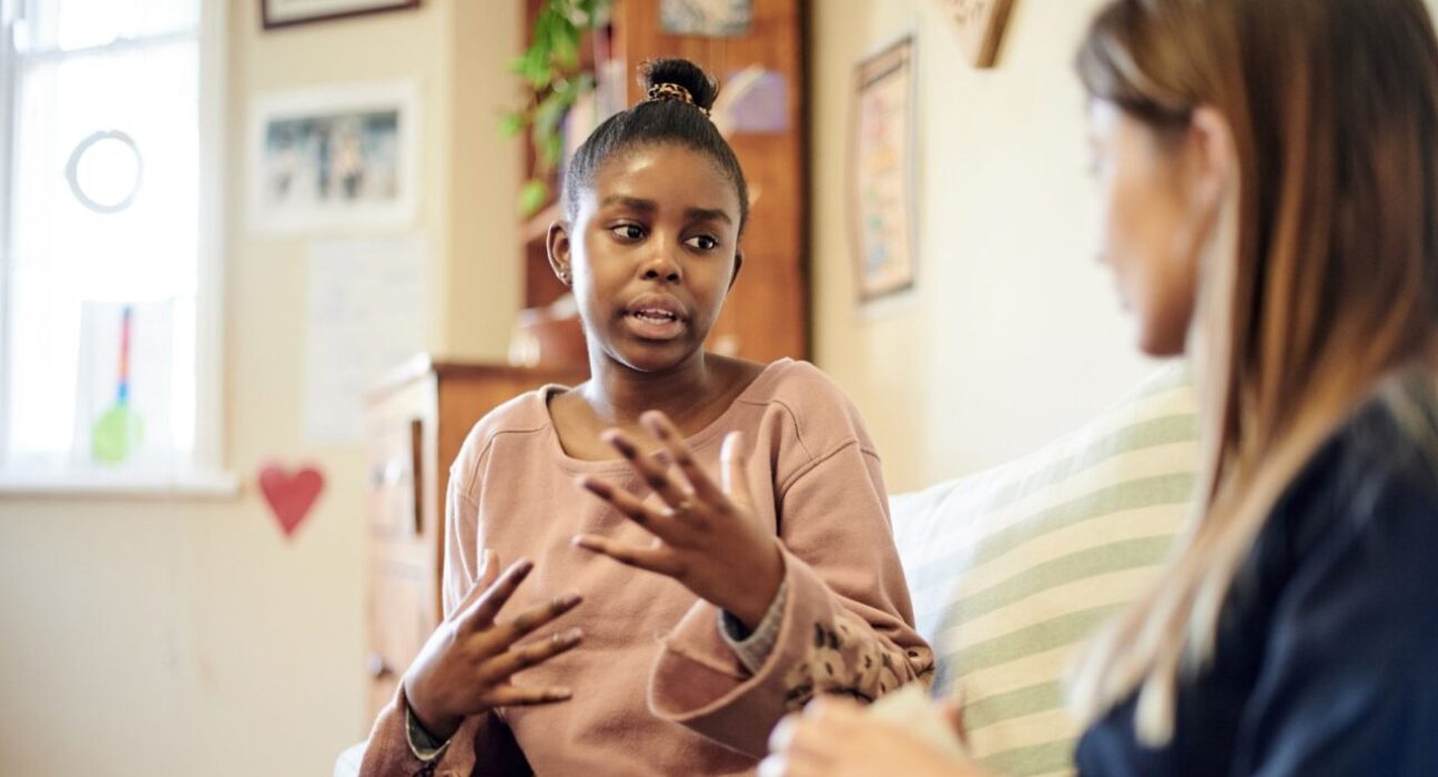 A woman is talking to a blurred woman on the couch. She is using hand gestures and making eye contact, she looks as though she is describing something difficult.