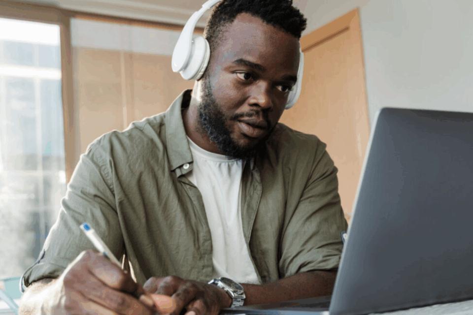 Man wearing headphones looking at a laptop screen whilst writing notes.