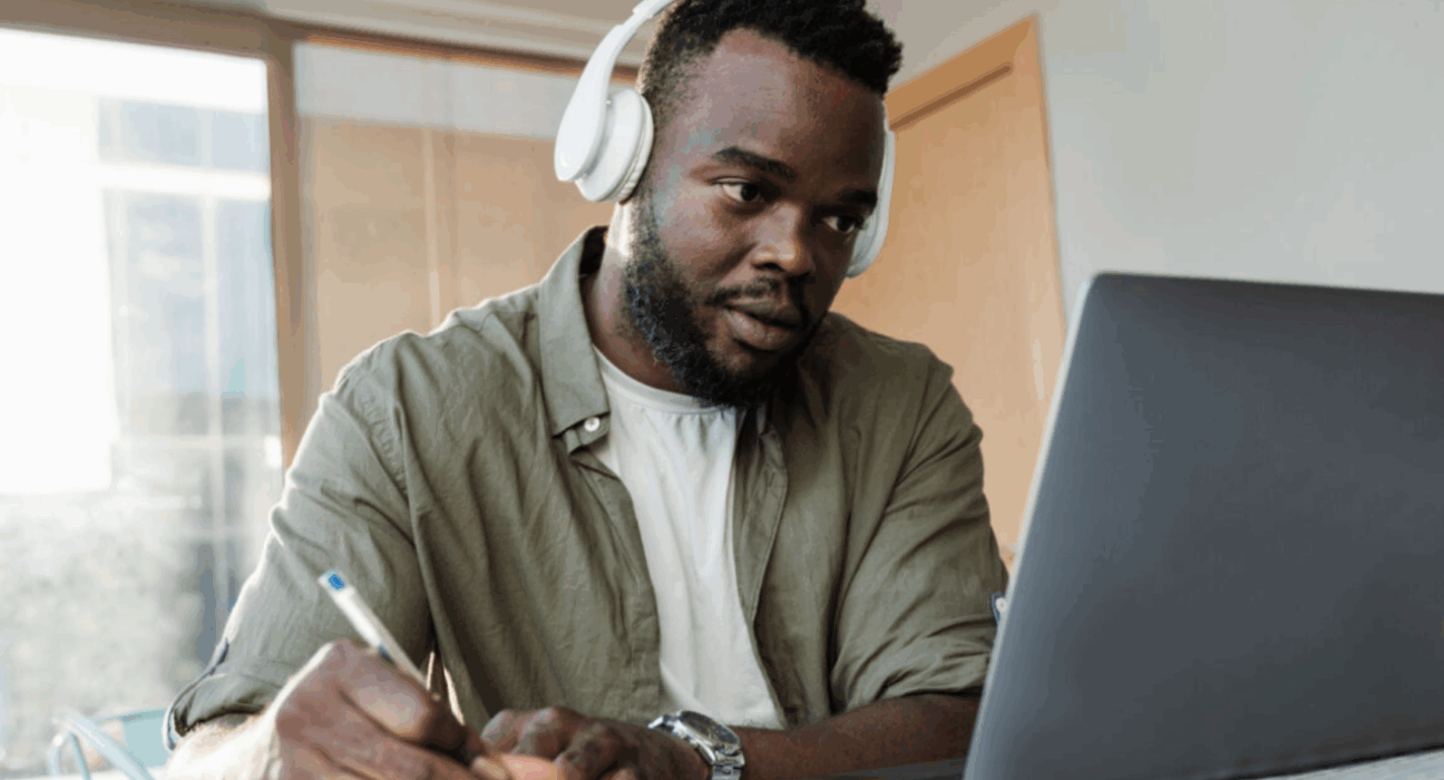 Man wearing headphones looking at a laptop screen whilst writing notes.