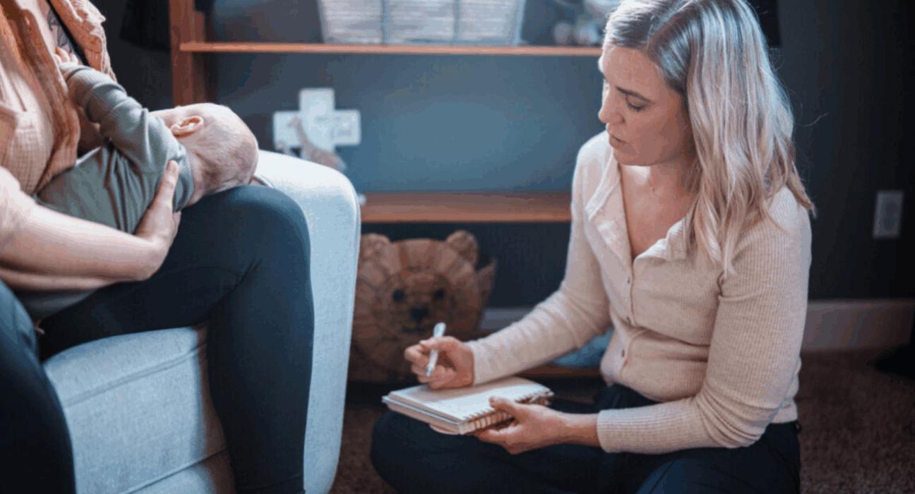 Lady sitting on the floor writing in a notepad, next to a lady sitting on the sofa holding a baby.
