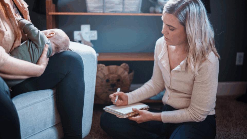 Lady sitting on the floor writing in a notepad, next to a lady sitting on the sofa holding a baby.